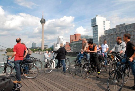 Menschen auf Fahrrädern an einem Ufer mit Blick auf den Fernsehturm in Düsseldorf.
