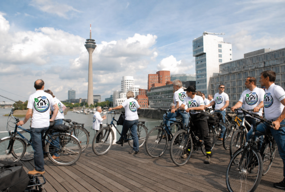 Gruppenausflug von Radfahrern am Fluss mit der Stadtsilhouette im Hintergrund.