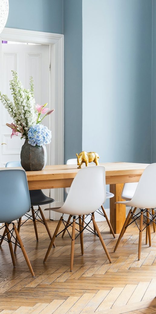 Heller Essbereich mit Holztablett, Stühlen in verschiedenen Farben und Blumenarrangement.